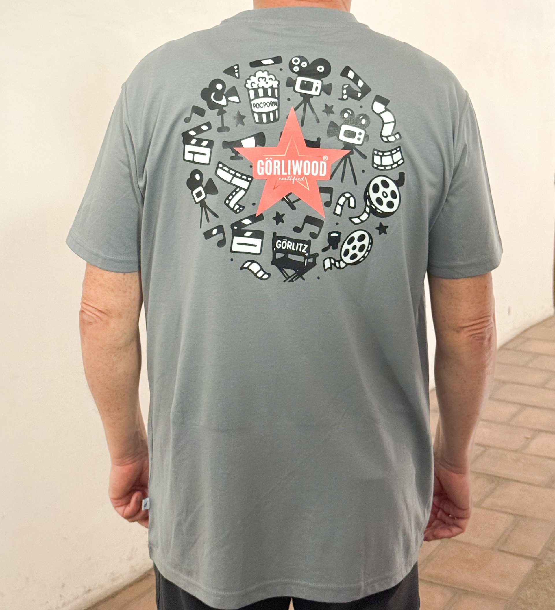 Auf der Rückseite des grauen, kurzärmligen Herren Shirts ist der Görliwood Stern zu sehen und kreisförmig dahinter in schwarz weiß verschiedene Motive aus dem Bereich Film.