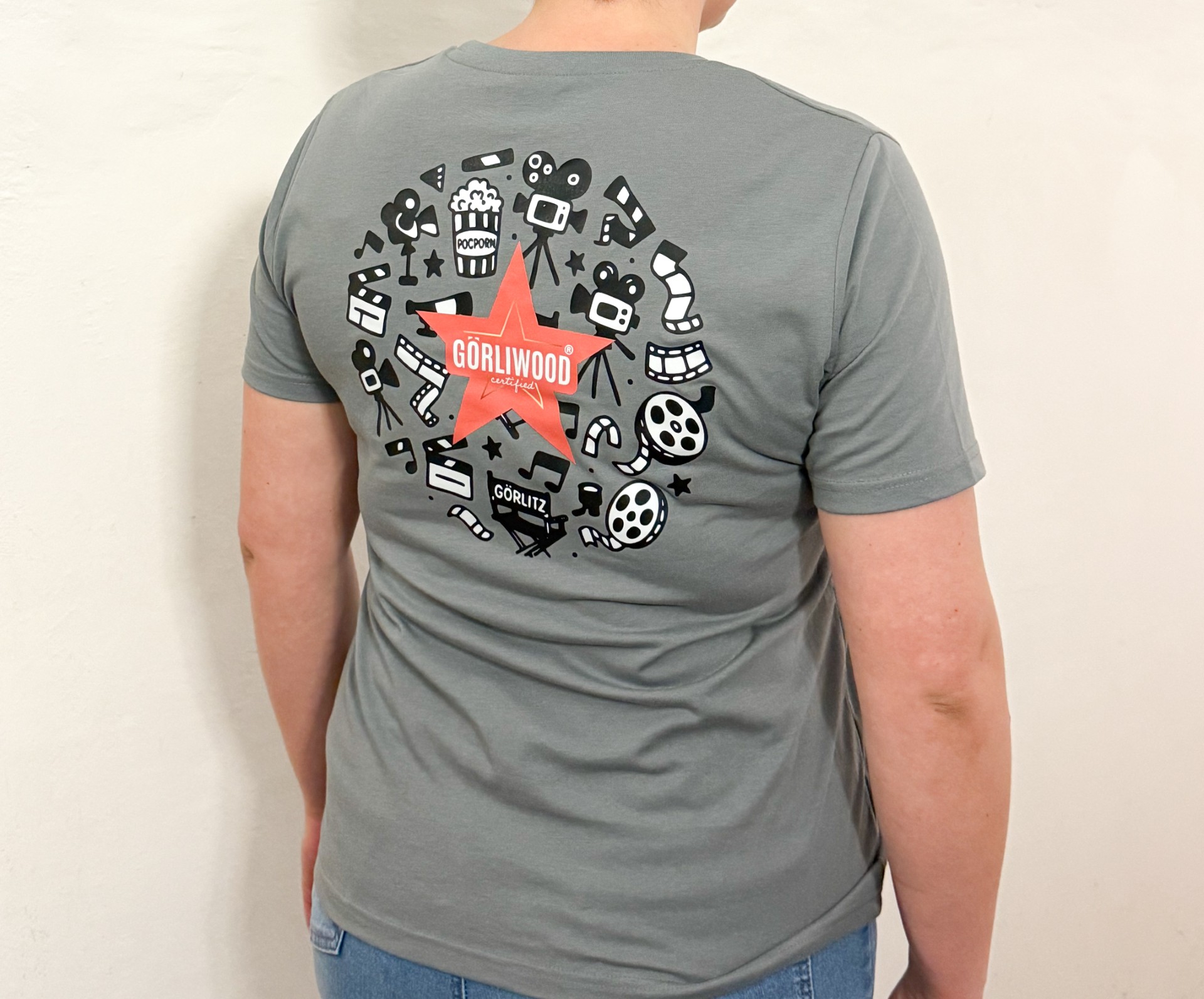 Auf der Rückseite des grauen, kurzärmligen Damen Shirts ist der Görliwood Stern zu sehen und kreisförmig dahinter in schwarz weiß verschiedene Motive aus dem Bereich Film.