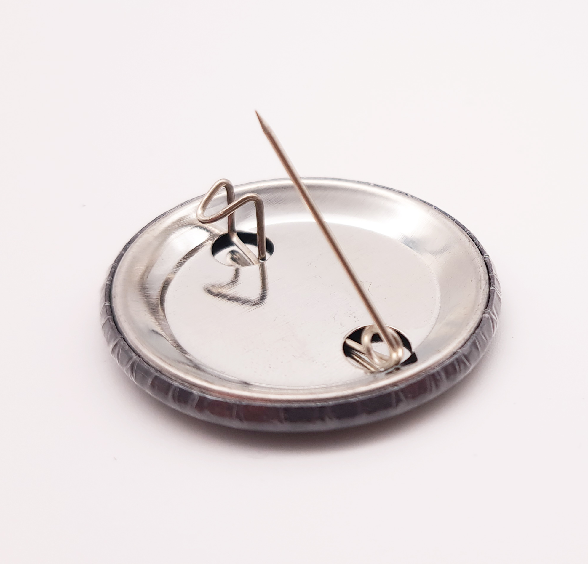 Der Button liegt nun mit der Bildseite auf dem Tisch und man sieht dass auf der Rückseite eine Befestigungsmöglichkeit mit der Nadel möglich ist. Diese ist auf dem Foto offen.