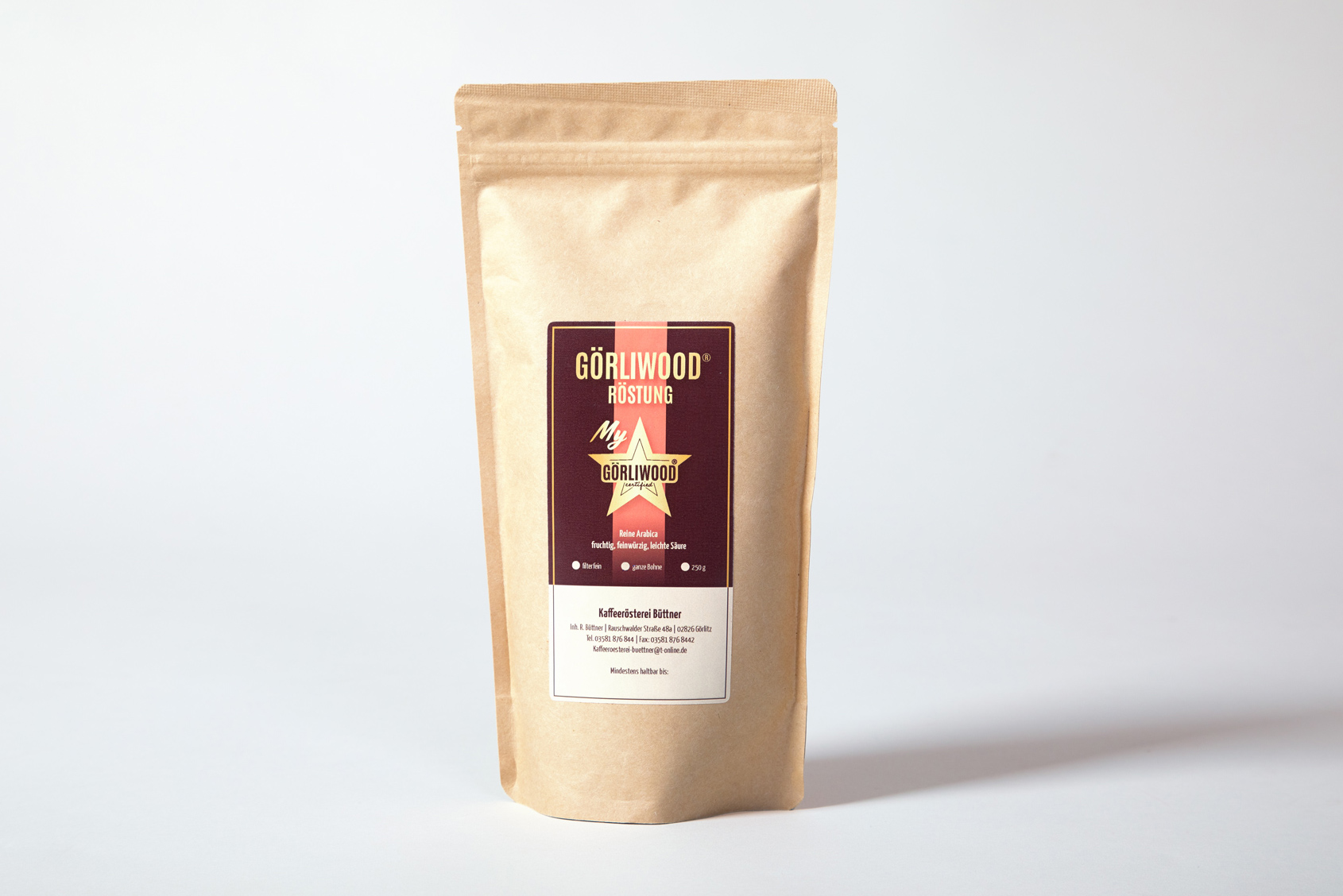 Görliwood-Kaffee von Vorne fotografiert. Die baige Kaffee-Tüte steht verschlossen mittig auf weißem Untergrund. Auf dem Etikett steht Görliwood Röstung und das Görliwood-Logo ist zu sehen. Der Hintergrund ist bräunlich und hat mittig einen roten Streifen.