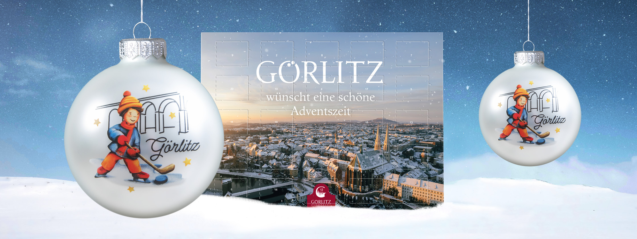 Der Schokoladenadventskalender  mit dem Foto von Görlitz im Winter aus der Vogelperspektive steht mittig im künstlichen Schnee. Rechts und links davon ist die Weihnachtsbaumkugel mit dem Motiv von 2025 zu sehen. Ein Junge beim Hockey spielen. Im Hintergru