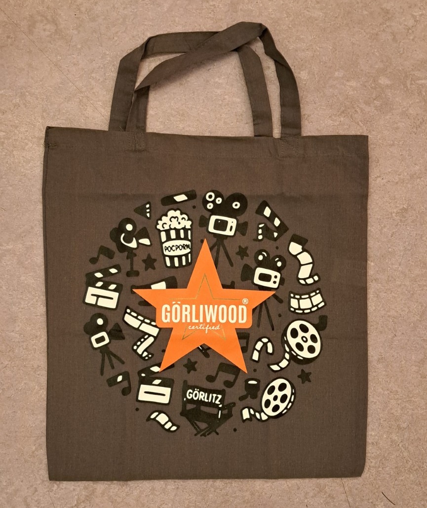 Der graue Stoffbeutel hat einen kurzen Henkel. Mittig ist der fünfzackige, orangefarbene Görliwoodstern zu sehen. Dahinter sind kreisförmig verschiedene Motive aus dem Bereich Film zu erkennen.