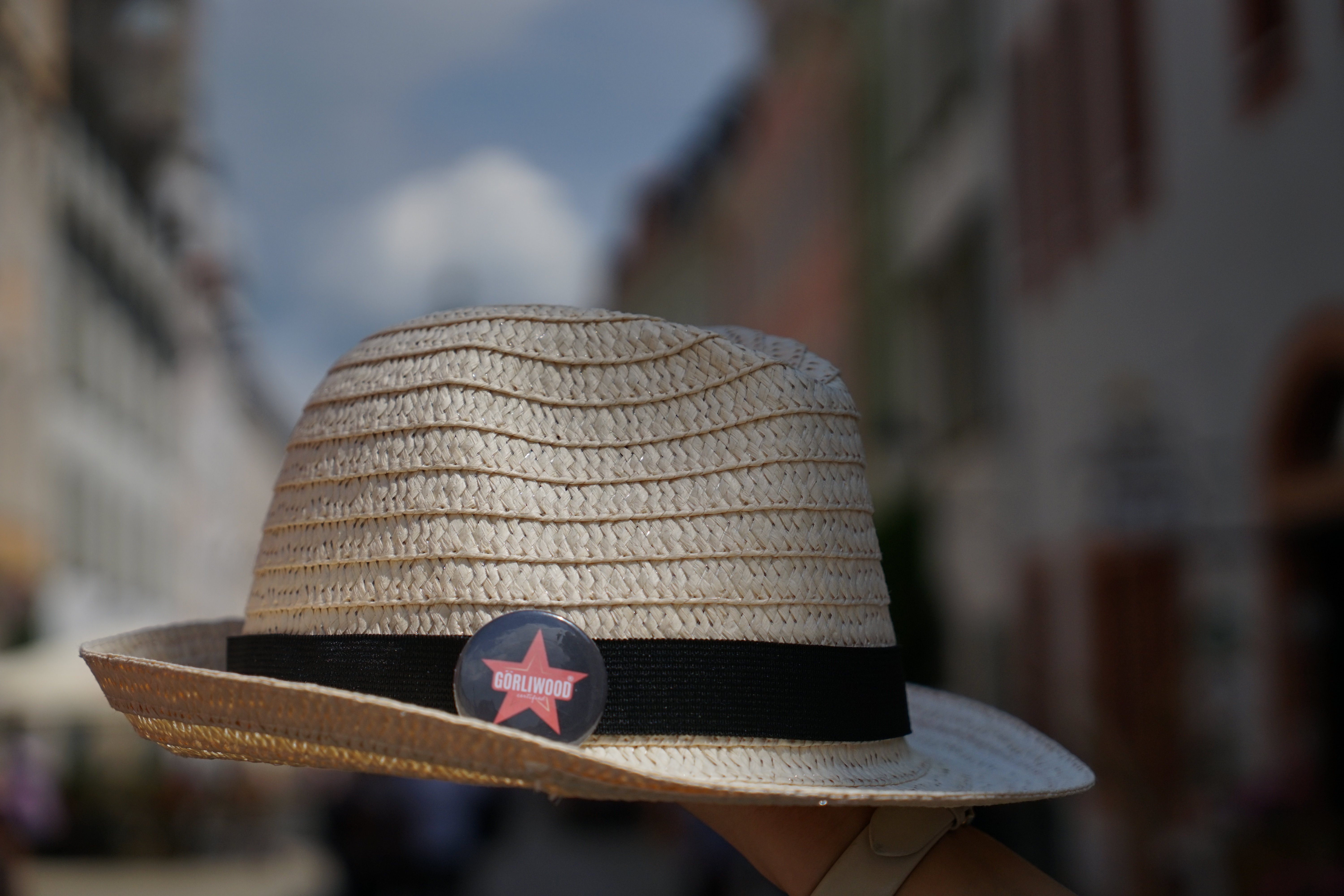 Der Strohhut mit einem schwarzen Band und dem Görliwood-Button wird hochgehalten. Im Hintergrund ist unscharf die Altstadt von Görlitz zu erahnen.