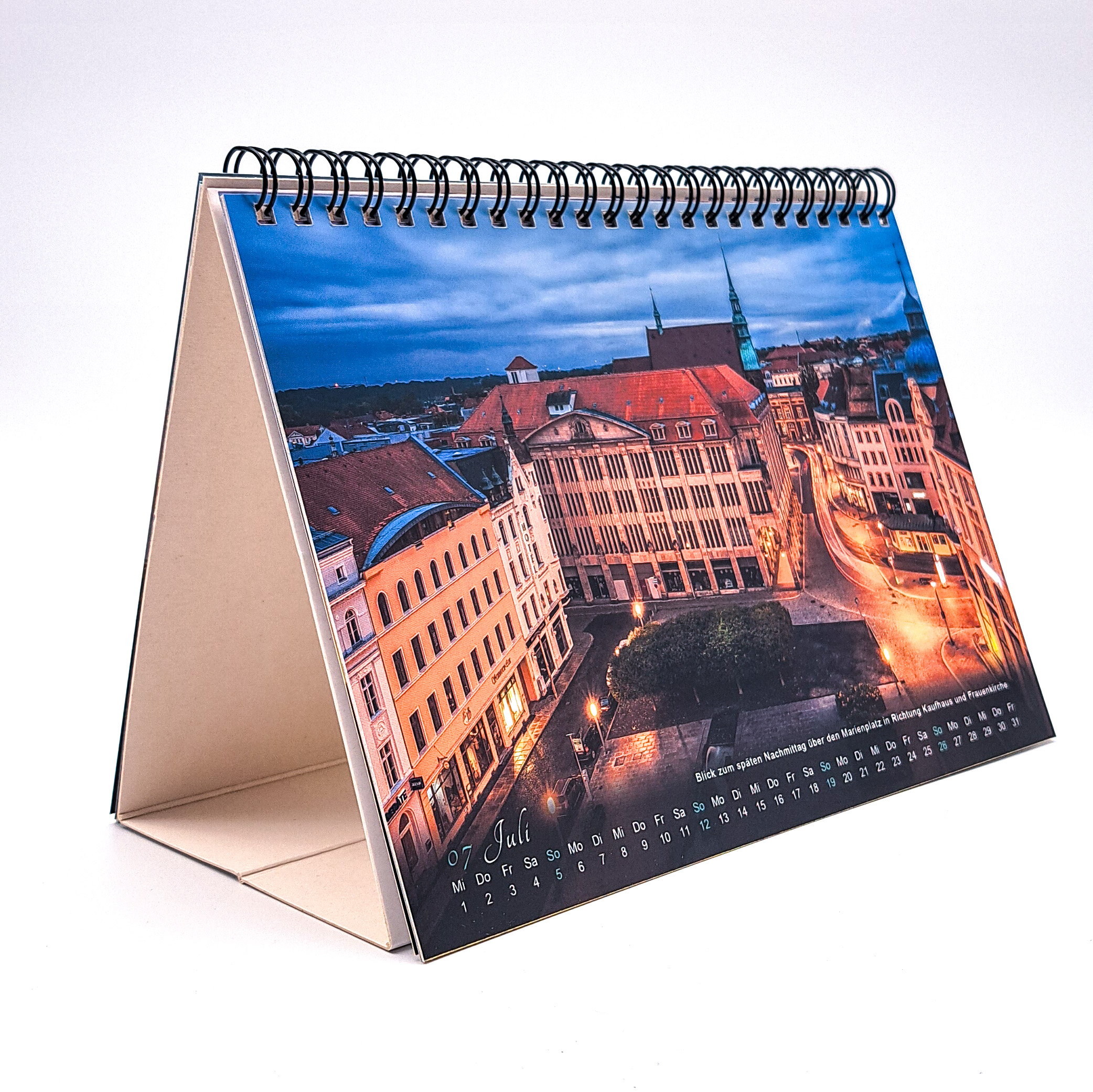 Der Tischkalender ist auf die Juli Ansicht geblättert. Zu sehen ist der Marienplatz mit dem Jugendstilkaufhaus und der Turmspitze der Frauenkirche bei stimmungsvollen Abendlicht. Am unteren Rand ist der Kalender in 2 Zeilen zu sehen. In der Oberen Zeile d