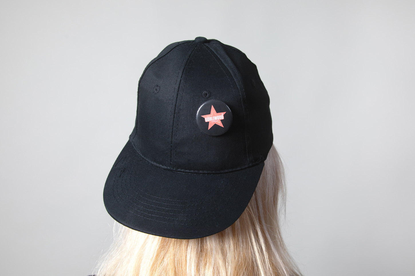 Görliwood-Cap Eine Person mit blonden, langen Haaren trägt ein schwarzes Cap mit dem Sonnenschild nach hinten. Über dem Sonnenschild ist der Görliwood-Button befestigt.