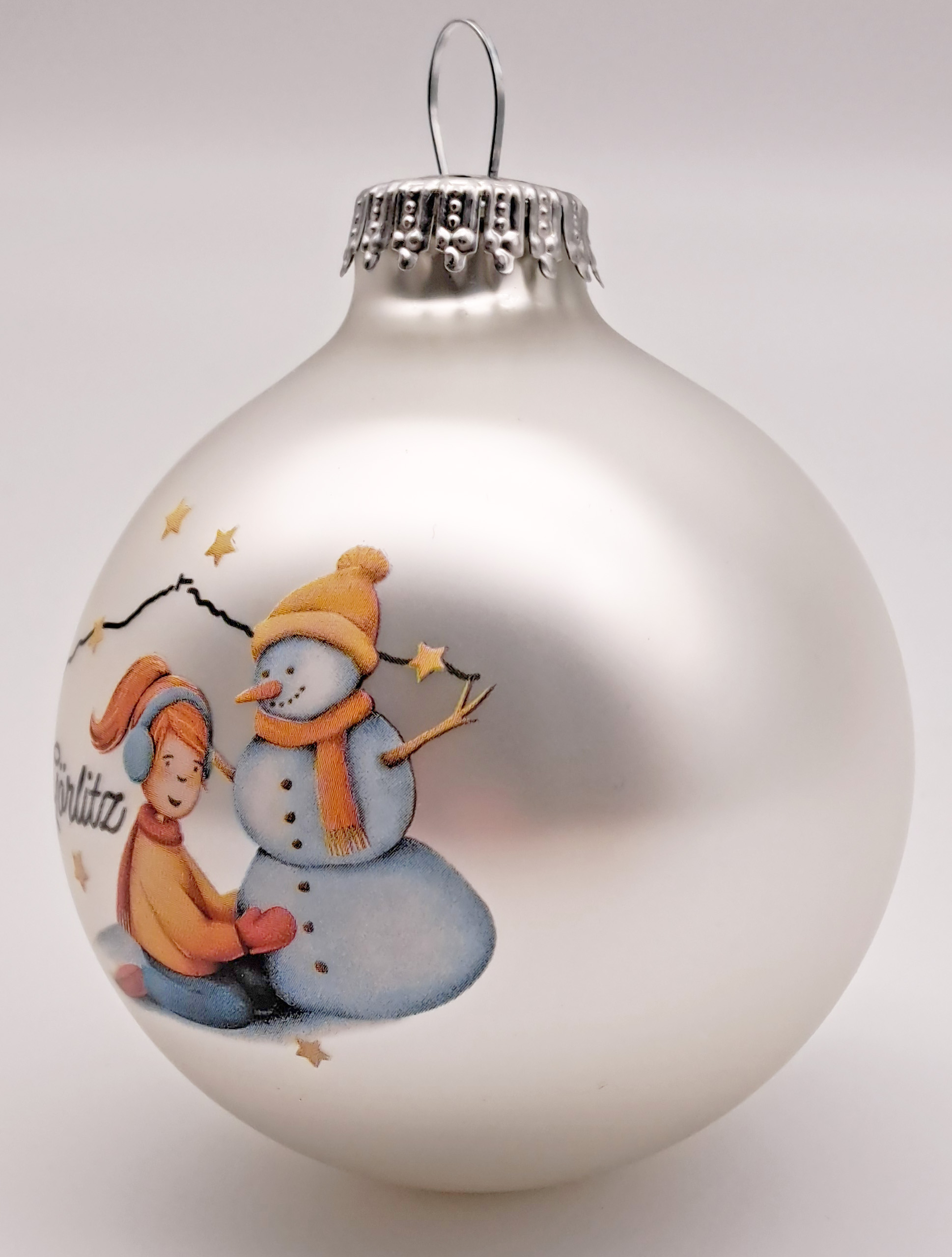 Man sieht eine Weihnachtskugel mit silberner Aufhängung, die Kugel ist in einem matten Silber und als gezeichnetes Motiv sieht man ein knieendes Mädchen, mit roten Haaren, Ohrwärmern rotem Schal und Handschuhen in gelber Jacke, die einen Schneemann baut. 