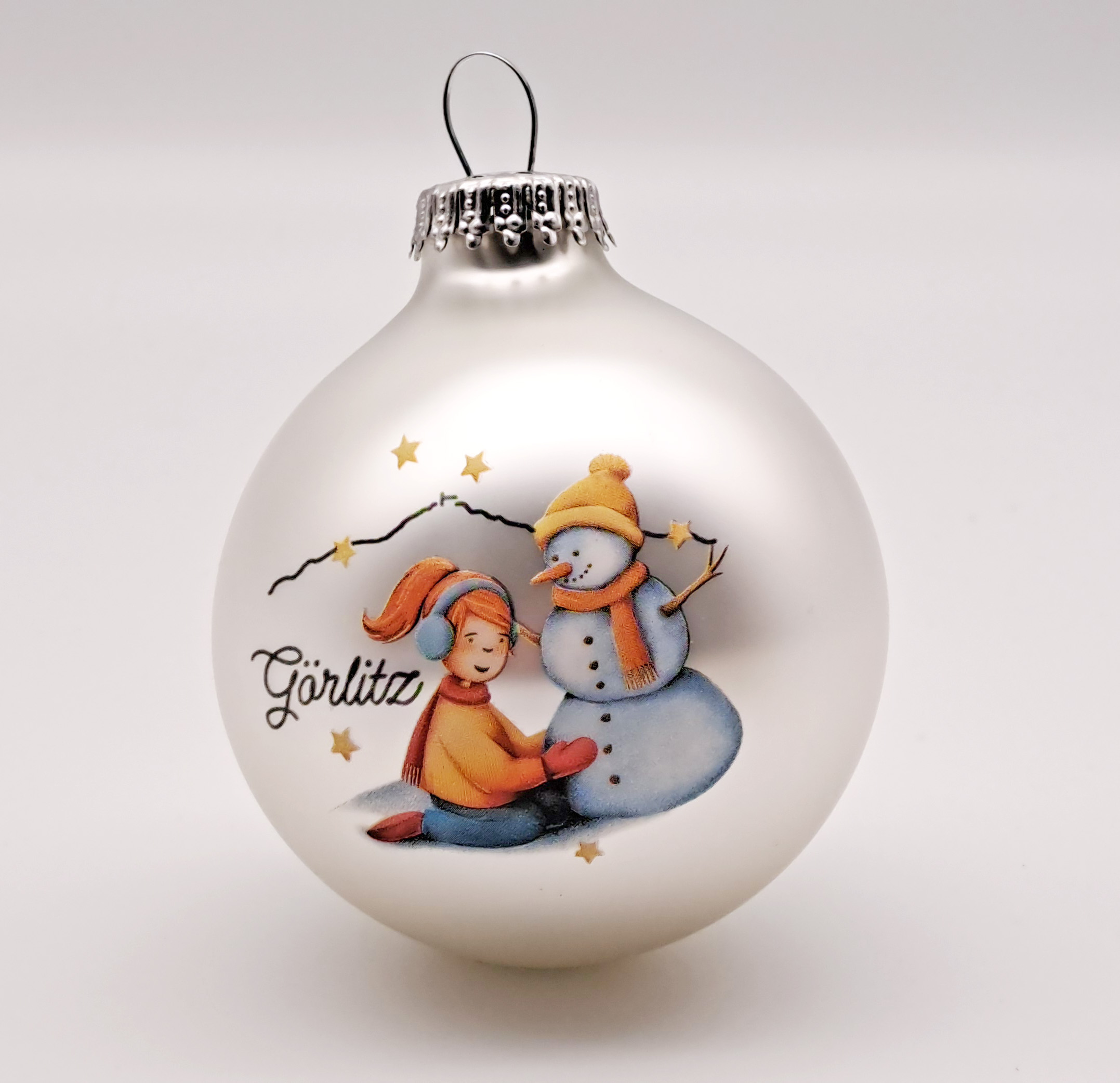 Man sieht eine Weihnachtskugel mit silberner Aufhängung, die Kugel ist in einem matten Silber und als gezeichnetes Motiv sieht man ein knieendes Mädchen, mit roten Haaren, Ohrwärmern rotem Schal und Handschuhen in gelber Jacke, die einen Schneemann baut. 