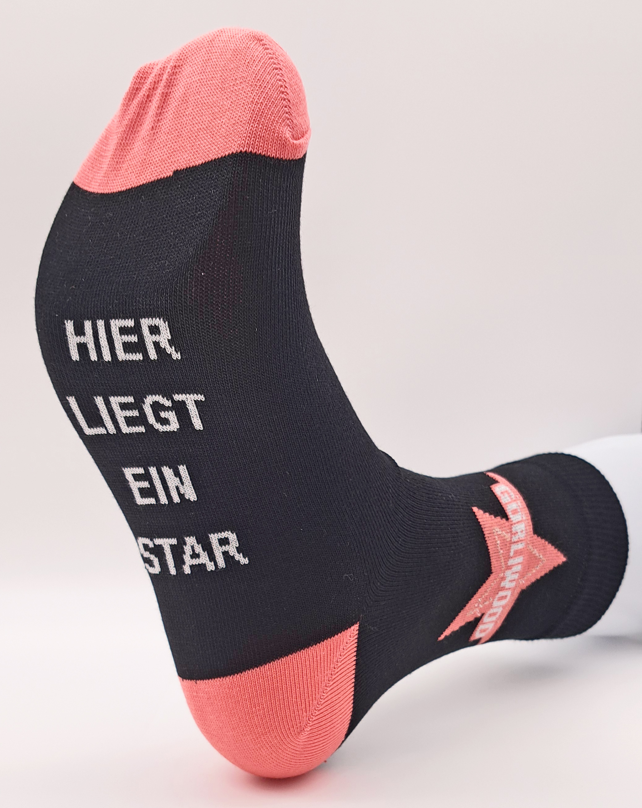 Eine schwarze Socke, die einem Fußmodel angezogen wurde. Bei dieser Socke sind die Zehen und die Ferse jedoch in einem Orange-Ton. Seitlich am Strumpf ist in dem selben Ton ein Stern, darin steht GÖRLIWOOD. Auf der Fußsohle steht in großen, weißen Buchsta
