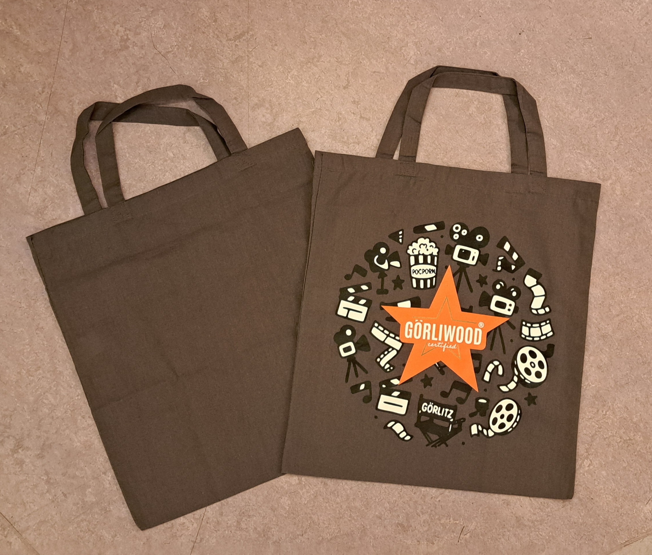 Zwei Tragetaschen sind etwas versetzt übereinander gelegt. links ist nur der graue Stoffbeutel zu sehen. Rechts ist mittig der fünfzackige Görliwood Stern zu sehen und kreisrund  in schwarz weiß verschiedene Motive aus dem Bereich Film. Die Taschen haben