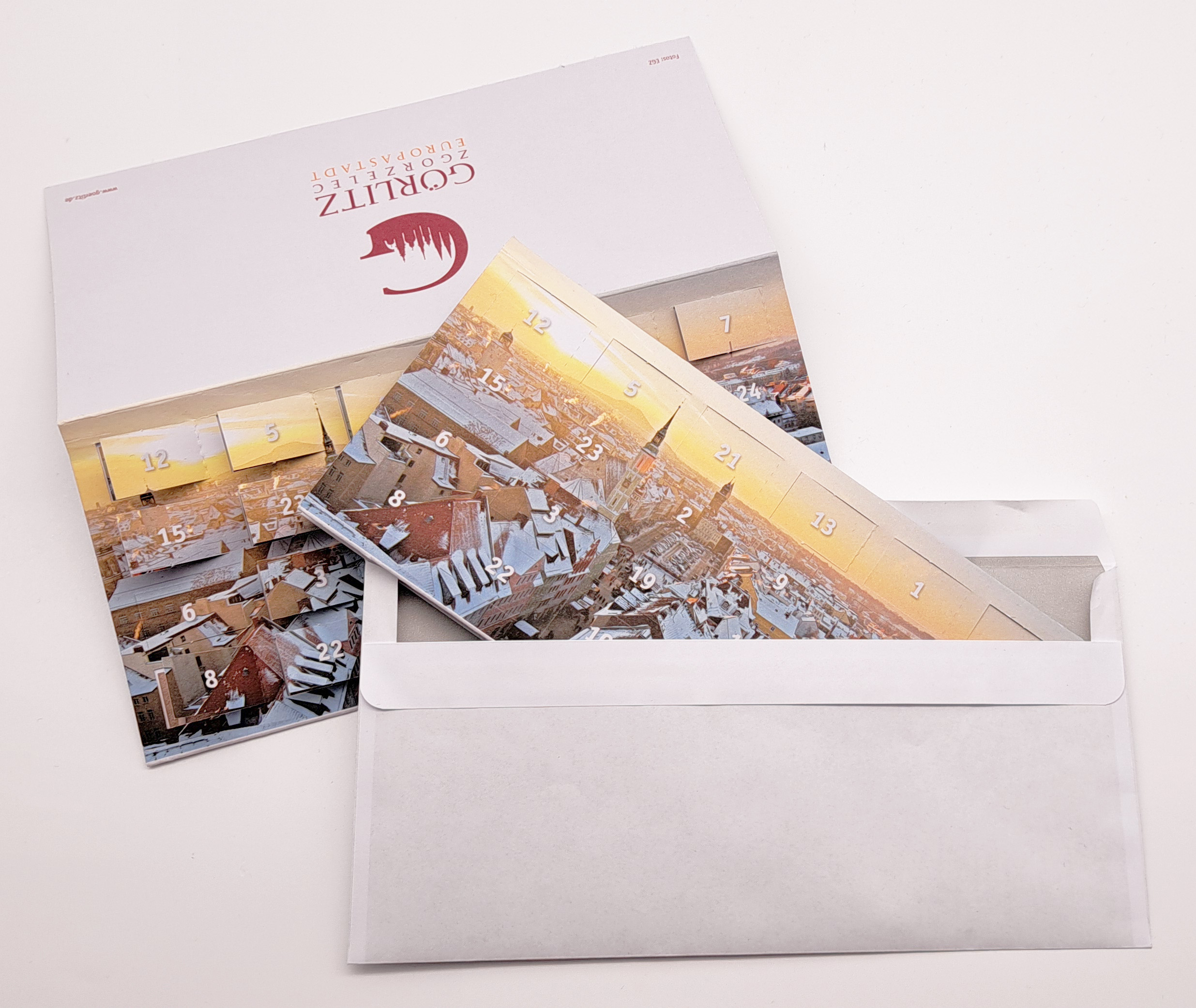 Adventskalender-Klappkarte im Kuvert im Vordergrund ist die Adventskalenderklappkarte halb in den Umschlag gesteckt. Im Hintergrund liegt eine aufgeklappte Adventskaleder-Klappkarte, bei der teilweise auch schon die Türchen geöffnet sind - man erkennt jedoch nicht, was dahinter ist.