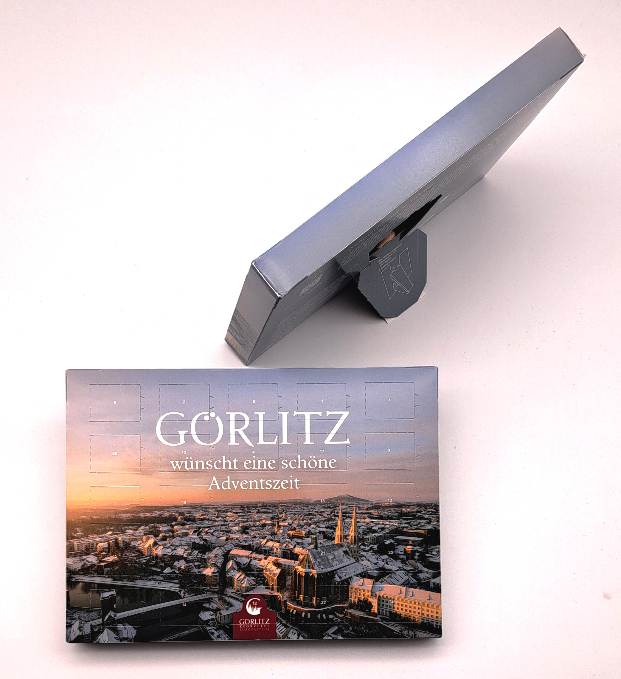 Der Adventskalender von 2025 hat auf der Titelseite ein winterliches Stadtpanorama bei Sonnenaufgang aus der Vogelperspektive. Hier sieht man diese Titelseite und oben rechts davon sieht man solch einen Kalender von oben, welcher auf der Rückseite den Tis