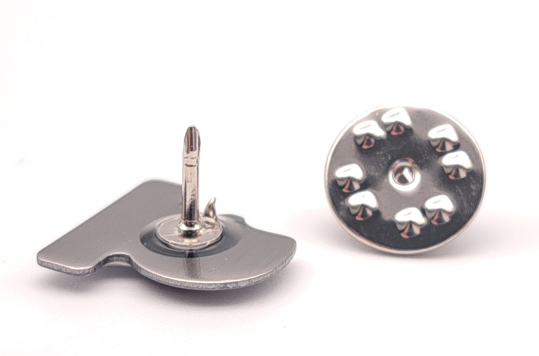 links liegt der Pin mit dem Motiv nach unten. Man erkennt die Nadel auf der Rückseite des Pins. Rechts daneben ist der Stecker dazu zu sehen. 