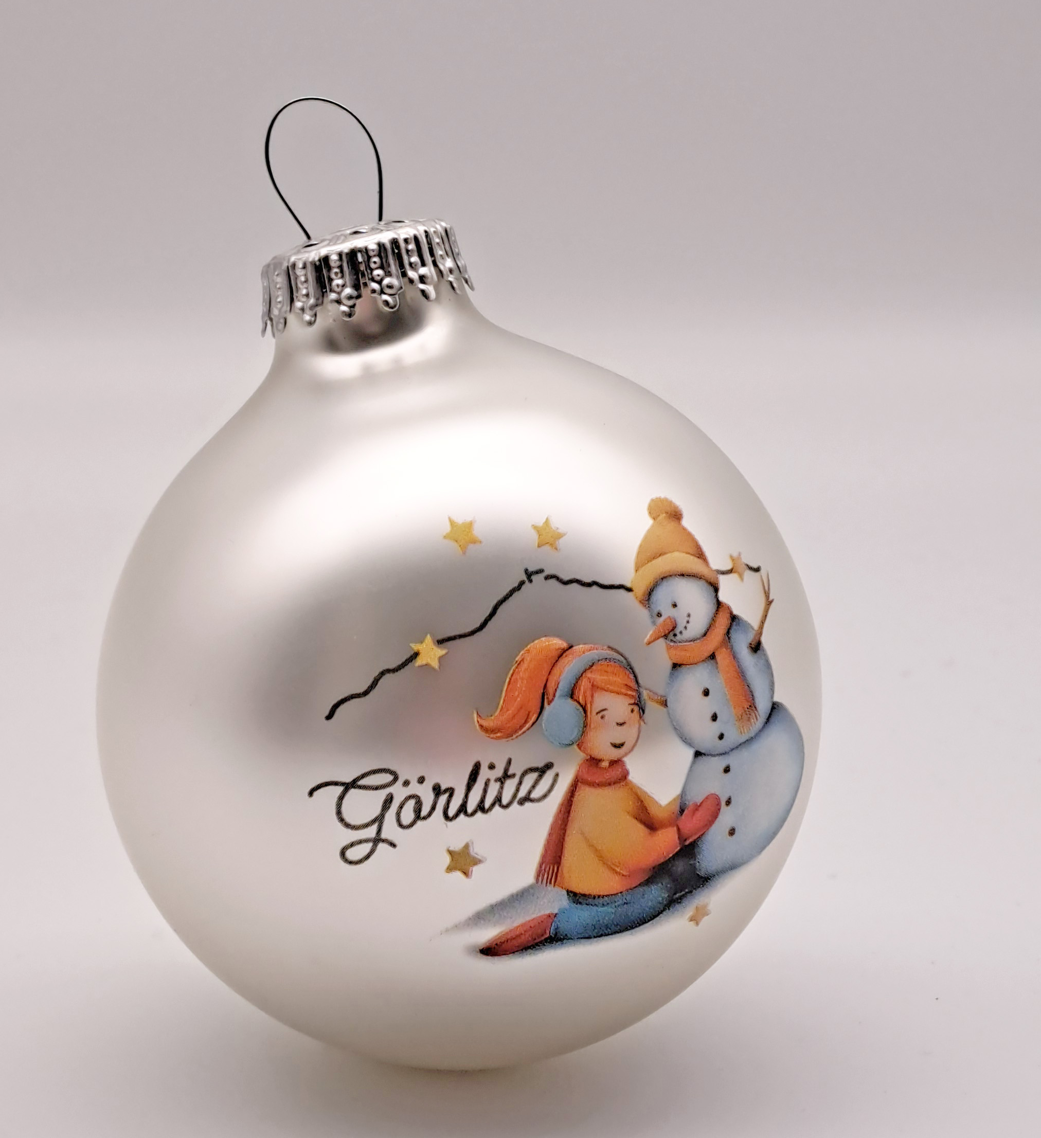 Man sieht eine Weihnachtskugel mit silberner Aufhängung, die Kugel ist in einem matten Silber und als gezeichnetes Motiv sieht man ein knieendes Mädchen, mit roten Haaren, Ohrwärmern rotem Schal und Handschuhen in gelber Jacke, die einen Schneemann baut. 