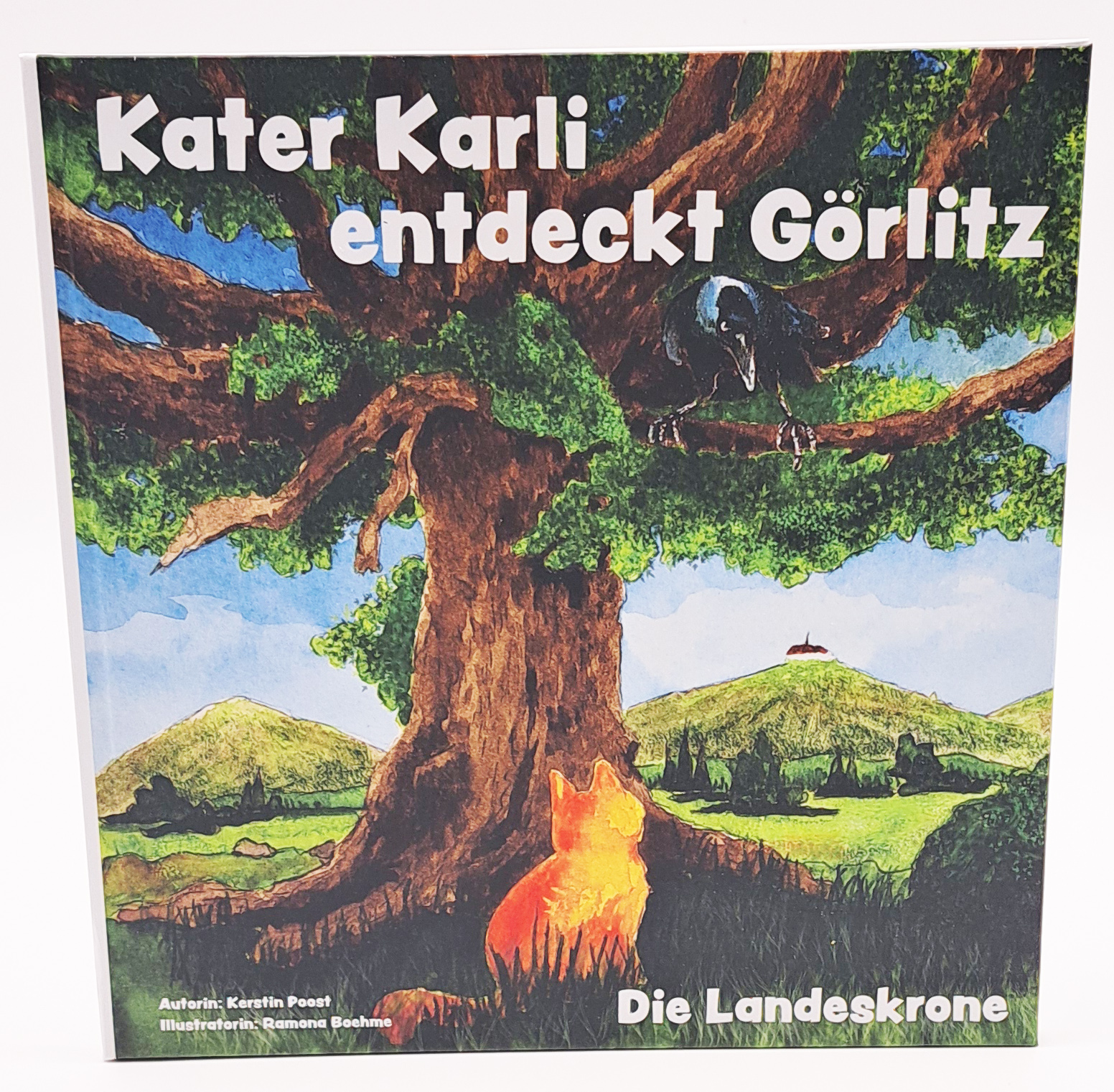 Auf der Titelseite steht in weiß Kater Karli entdeckt Görlitz Die Landeskrone. Man erkennt im Hintergrund die Landeskrone. Kater Karli sitz mit dem Rücken zum Leser vor einem großen Baum. In der Baumkrone sitzt ein Rabe.